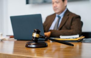 Image of a judge gavel on a table with a person sitting at the back with laptop